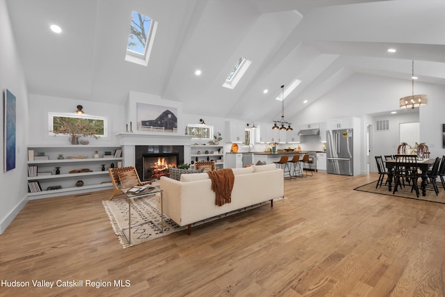 living room featuring a skylight, high vaulted ceiling, a glass covered fireplace, a chandelier, and light wood-type flooring