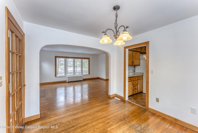 empty room featuring arched walkways, baseboards, radiator heating unit, light wood-style flooring, and an inviting chandelier