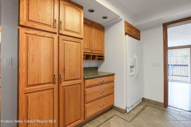kitchen featuring dark stone countertops, light tile patterned flooring, baseboards, brown cabinets, and white fridge with ice dispenser