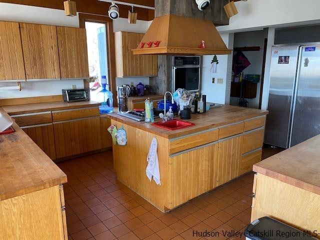 kitchen with custom range hood, sink, butcher block countertops, black oven, and stainless steel refrigerator