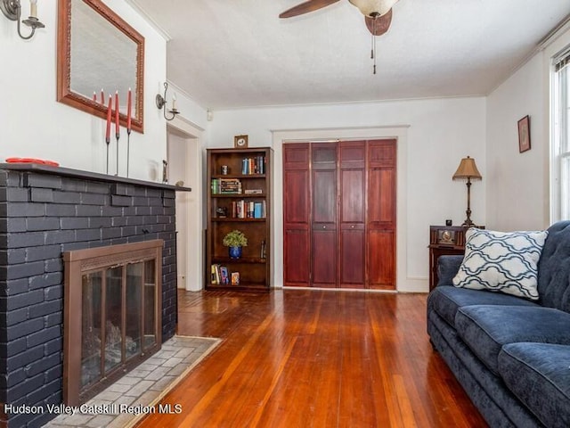 living room with dark wood-type flooring, ceiling fan, a brick fireplace, and crown molding