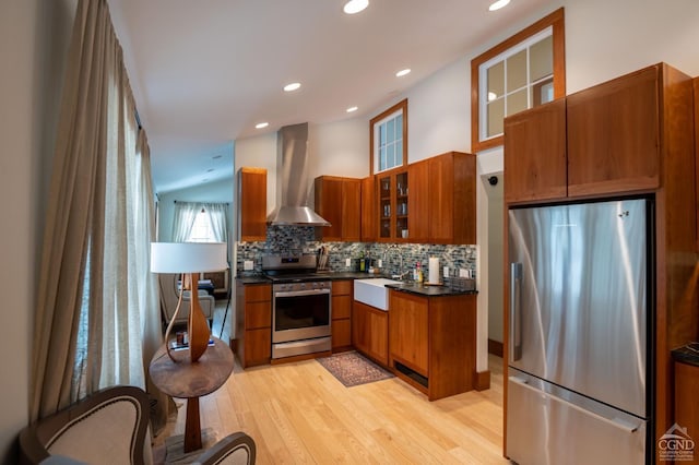 - kitchen featuring appliances with stainless steel finishes, wall chimney exhaust hood, brown cabinetry, light wood-type flooring, and dark countertops