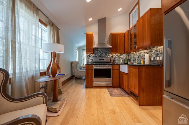 - kitchen with appliances with stainless steel finishes, wall chimney exhaust hood, brown cabinetry, light wood-type flooring, and decorative backsplash