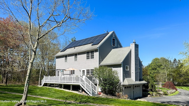 rear view of house featuring a lawn, a wooden deck, a chimney, and roof mounted solar panels