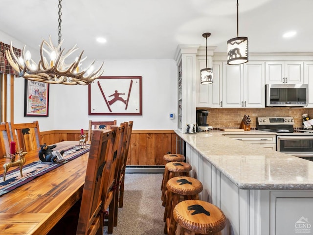 kitchen featuring stainless steel appliances, a wainscoted wall, baseboard heating, white cabinets, and decorative light fixtures