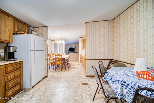 kitchen featuring brown cabinetry, wallpapered walls, freestanding refrigerator, light countertops, and a notable chandelier