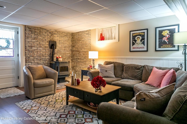 living area featuring brick wall, a drop ceiling, wainscoting, a wood stove, and wood finished floors