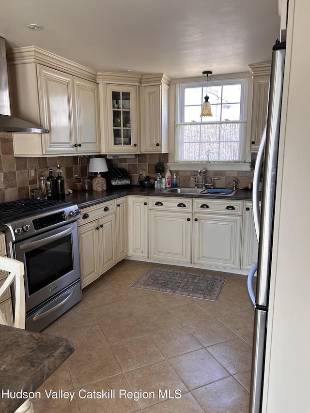 kitchen featuring dark countertops, wall chimney range hood, cream cabinetry, stainless steel appliances, and a sink