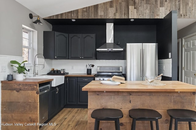 kitchen featuring appliances with stainless steel finishes, butcher block counters, wall chimney exhaust hood, lofted ceiling, and a sink