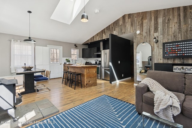living room featuring light wood-style floors, high vaulted ceiling, arched walkways, a skylight, and wooden walls