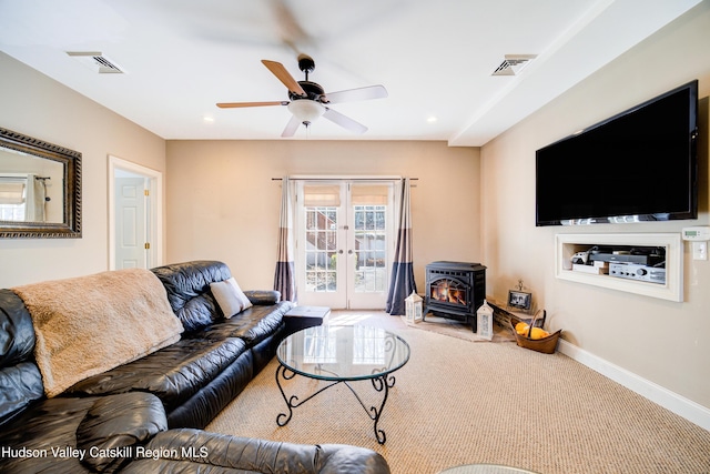 carpeted living room featuring visible vents, baseboards, a wood stove, a ceiling fan, and french doors