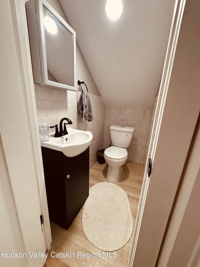 half bathroom with vaulted ceiling, toilet, vanity, tile walls, and wood finished floors