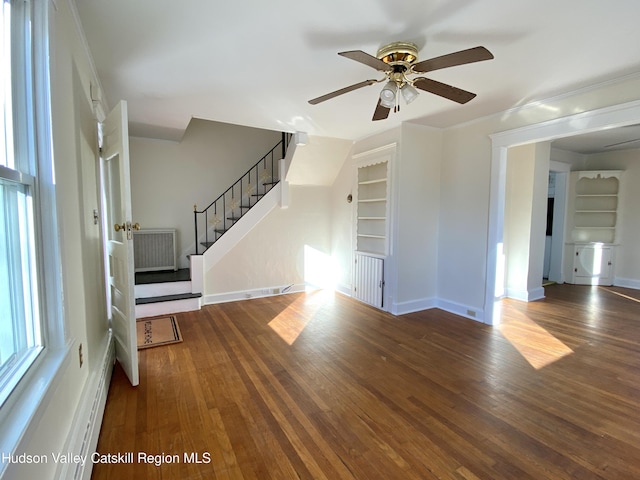 unfurnished living room with ceiling fan, built in features, dark wood-type flooring, and ornamental molding