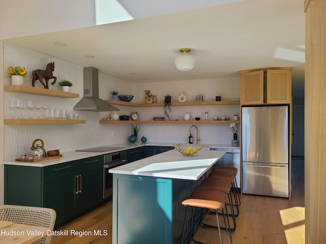 kitchen with a center island, wall chimney range hood, appliances with stainless steel finishes, light hardwood / wood-style floors, and a kitchen bar