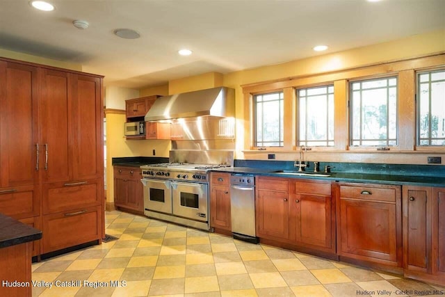 kitchen featuring a sink, dark countertops, wall chimney exhaust hood, brown cabinetry, and appliances with stainless steel finishes