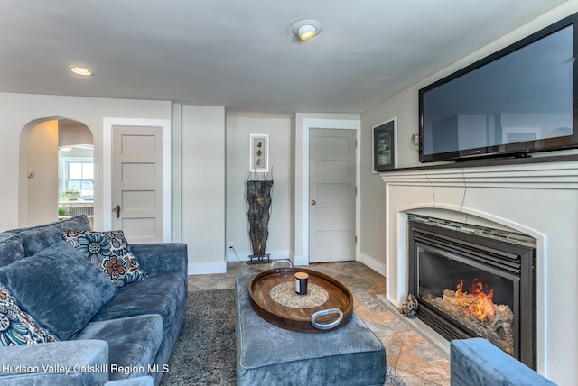 living room featuring arched walkways, stone tile floors, recessed lighting, a glass covered fireplace, and baseboards