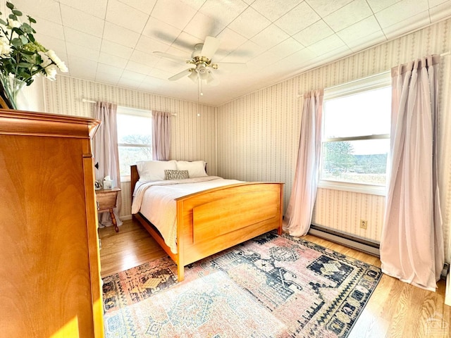 bedroom with baseboard heating, wood finished floors, wallpapered walls, and a ceiling fan