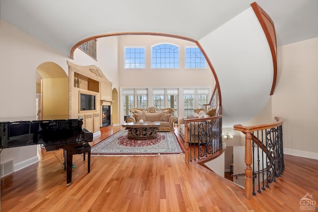 living area featuring wood finished floors, a towering ceiling, a glass covered fireplace, baseboards, and arched walkways