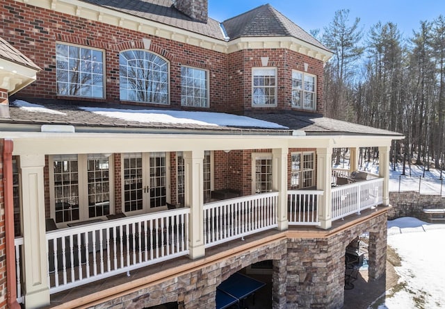 snow covered property with french doors, brick siding, a balcony, and a chimney