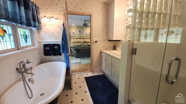 ensuite bathroom featuring wallpapered walls, a shower stall, a freestanding bath, a ceiling fan, and wainscoting