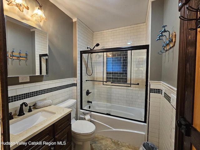 bathroom featuring toilet, vanity, tile walls, combined bath / shower with glass door, and wainscoting