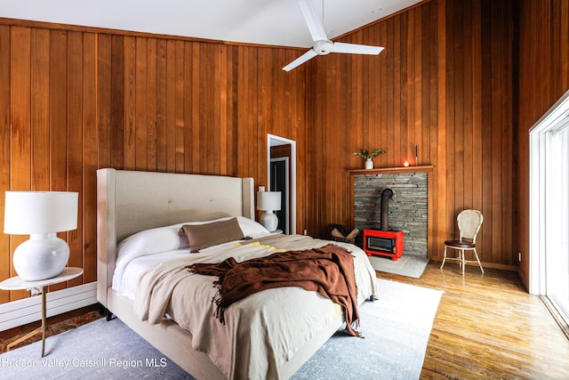 bedroom featuring baseboard heating, wooden walls, a wood stove, ceiling fan, and light hardwood / wood-style flooring