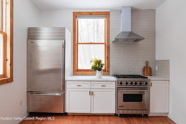 kitchen with premium appliances, decorative backsplash, white cabinets, light hardwood / wood-style flooring, and wall chimney exhaust hood