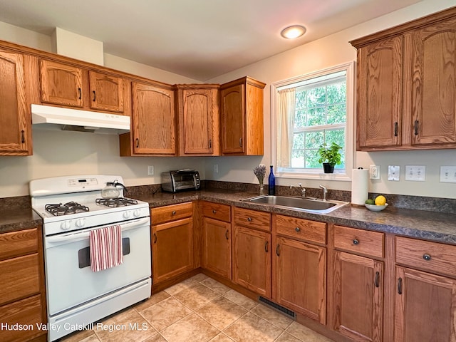 kitchen with brown cabinetry, dark countertops, white range with gas cooktop, under cabinet range hood, and a sink