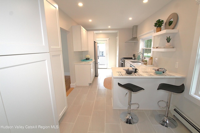 kitchen with a baseboard radiator, wall chimney range hood, stainless steel appliances, a kitchen bar, and white cabinets