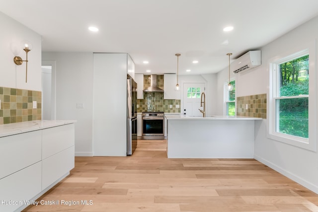 kitchen featuring white cabinetry, wall chimney exhaust hood, hanging light fixtures, an AC wall unit, and appliances with stainless steel finishes