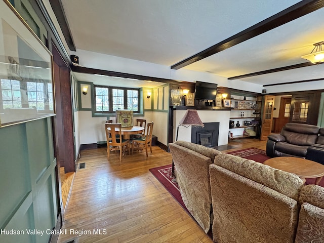 living area with visible vents, baseboards, a fireplace with flush hearth, beamed ceiling, and light wood-style floors