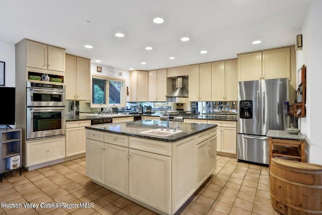 kitchen with cream cabinetry, stainless steel appliances, an island with sink, dark stone counters, and wall chimney exhaust hood