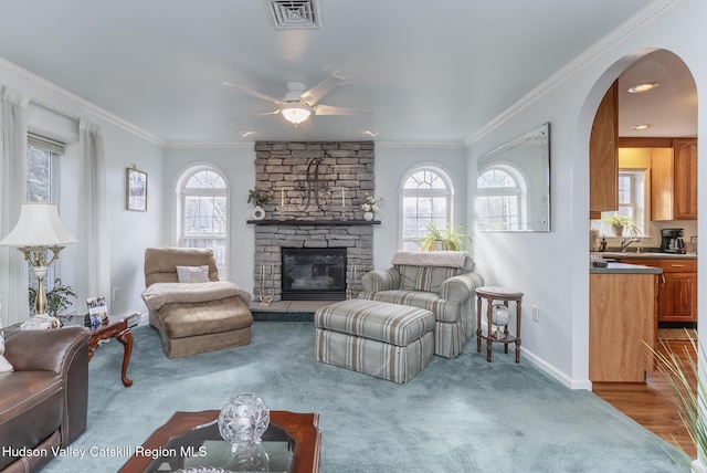 living room featuring a stone fireplace, crown molding, a ceiling fan, and visible vents