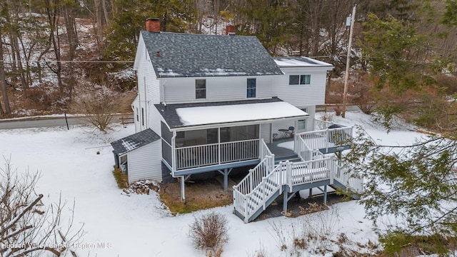 snow covered house with a sunroom, a chimney, roof with shingles, stairs, and a deck