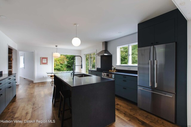 kitchen with sink, wall chimney exhaust hood, dark wood-type flooring, a kitchen island with sink, and appliances with stainless steel finishes