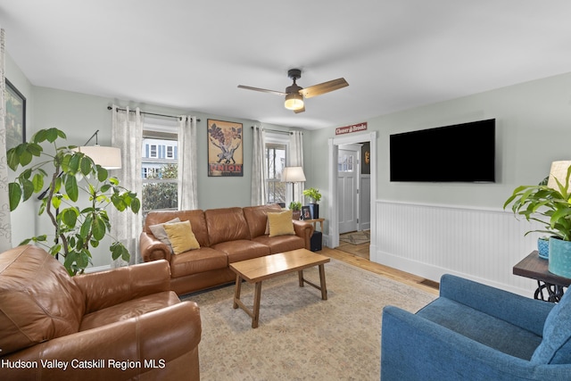 living area featuring light wood-type flooring, a ceiling fan, and wainscoting