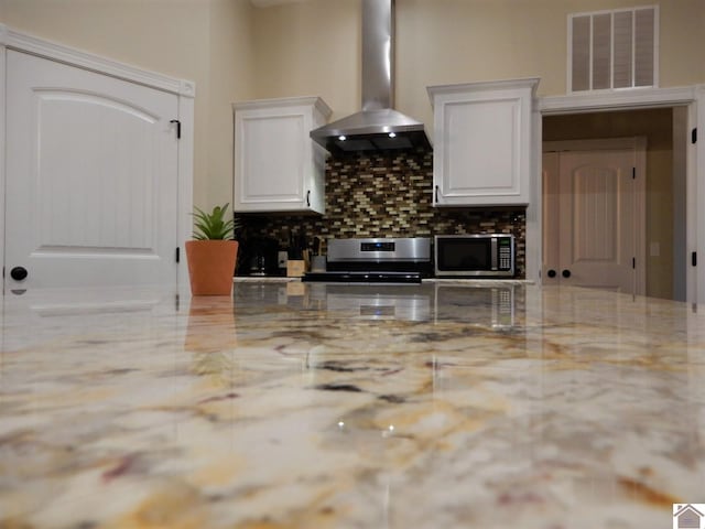 kitchen featuring wall chimney range hood, backsplash, range, light stone countertops, and white cabinets