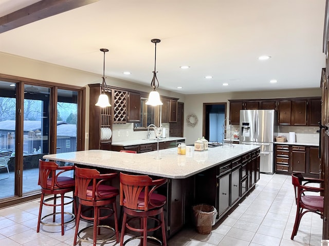 kitchen with an island with sink, light tile floors, light stone countertops, stainless steel fridge with ice dispenser, and hanging light fixtures