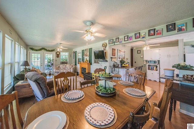 dining space featuring light wood-style flooring, a fireplace, ceiling fan, french doors, and a textured ceiling