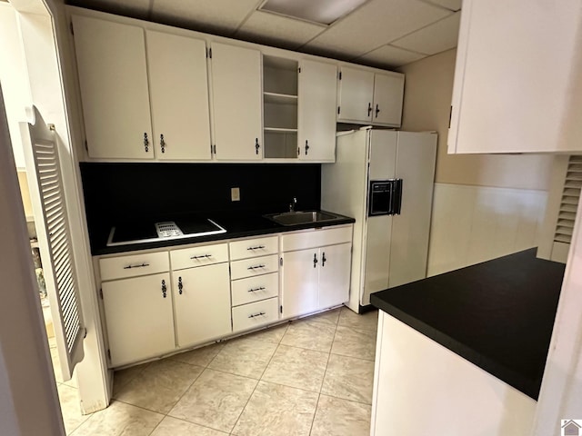 kitchen with white refrigerator with ice dispenser, open shelves, dark countertops, a paneled ceiling, and a sink