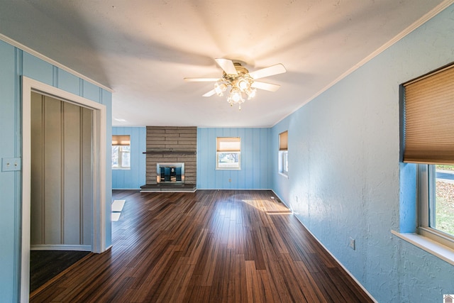 unfurnished living room featuring ceiling fan, wooden walls, dark wood-type flooring, crown molding, and a wood stove