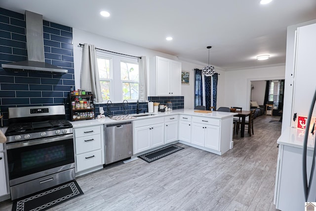 kitchen featuring wall chimney range hood, light hardwood / wood-style flooring, kitchen peninsula, decorative light fixtures, and appliances with stainless steel finishes