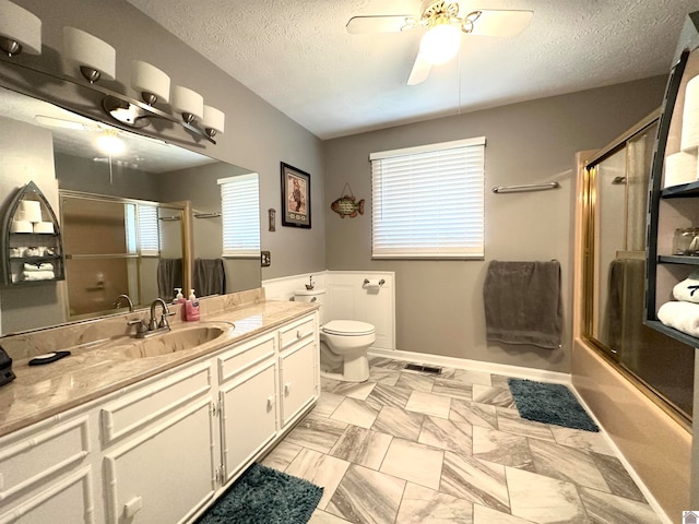 full bathroom with combined bath / shower with glass door, vanity, a textured ceiling, ceiling fan, and toilet