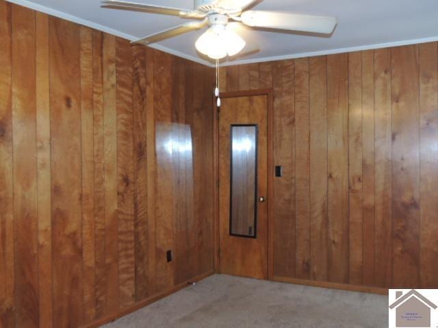 empty room with ceiling fan, wood walls, light colored carpet, and crown molding