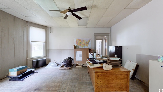 interior space featuring ceiling fan, radiator heating unit, a drop ceiling, wood walls, and hardwood / wood-style flooring