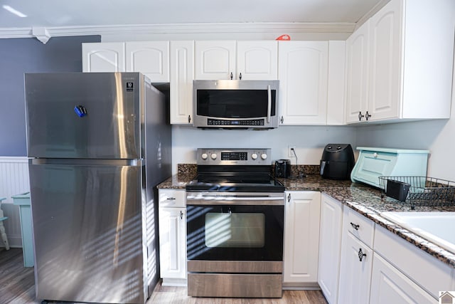 kitchen featuring appliances with stainless steel finishes, crown molding, dark stone countertops, white cabinets, and light hardwood / wood-style floors