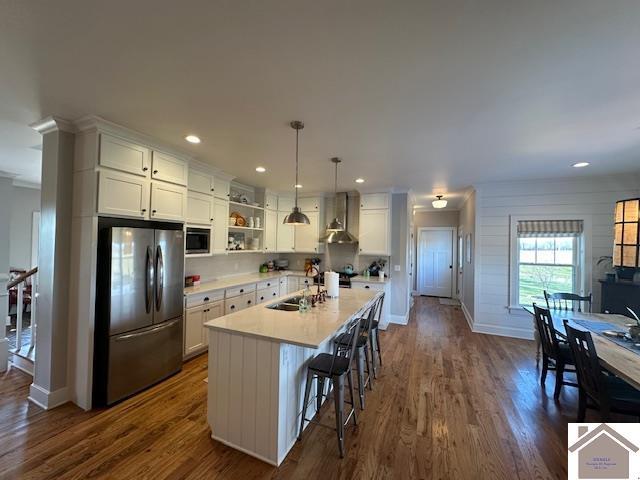 kitchen featuring a kitchen island with sink, stainless steel appliances, pendant lighting, wall chimney exhaust hood, and white cabinets