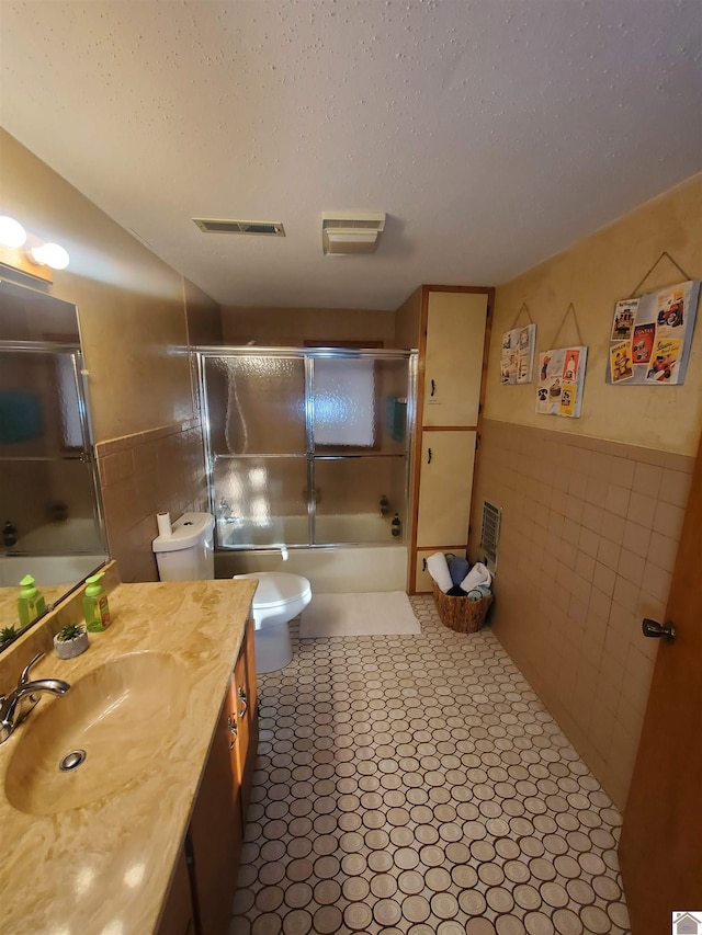 full bathroom with toilet, vanity, tile walls, bath / shower combo with glass door, and a textured ceiling