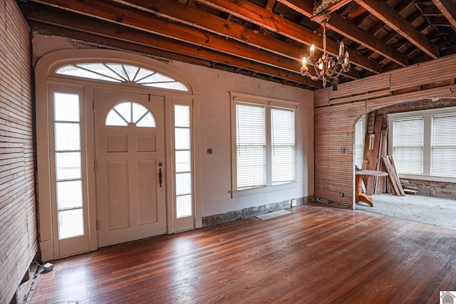 entrance foyer with brick wall, dark wood-type flooring, a chandelier, and beam ceiling