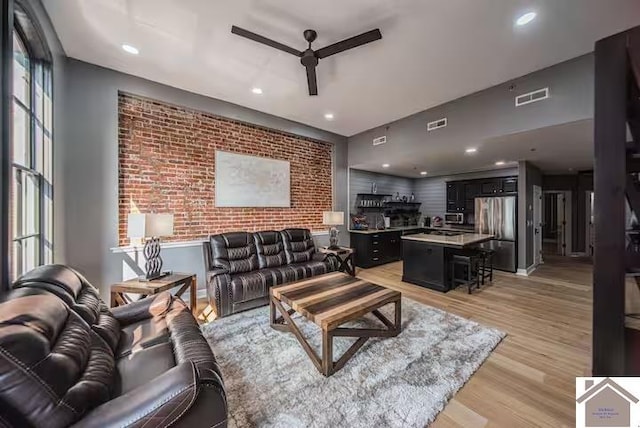 living area featuring light wood finished floors, visible vents, a ceiling fan, an accent wall, and brick wall